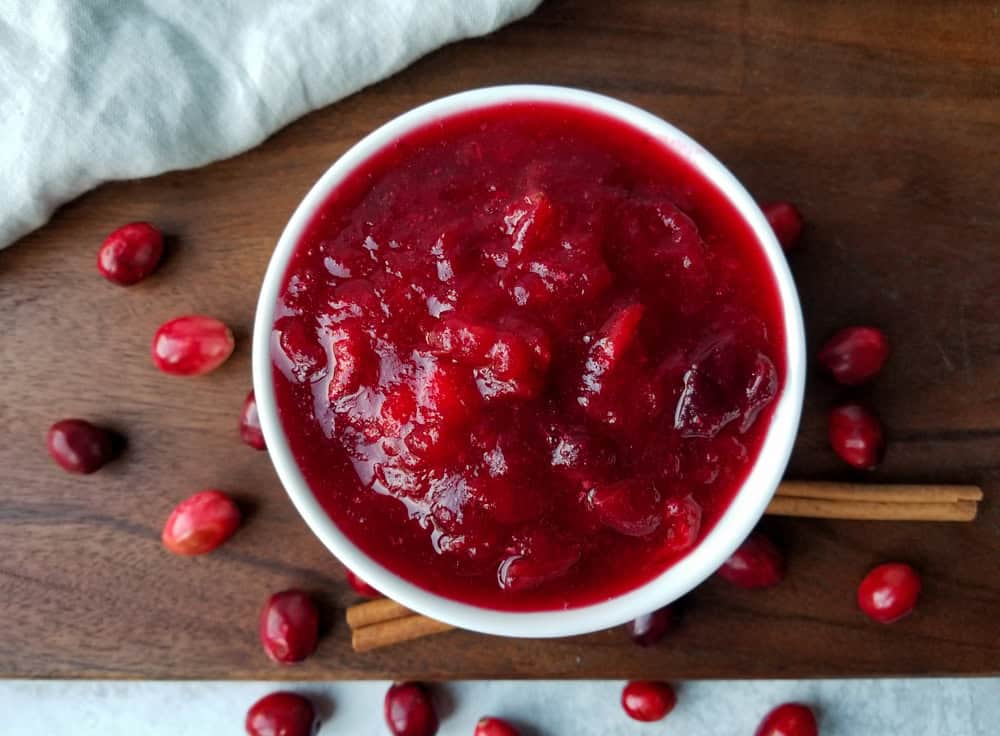 A round white bowl filled with thick, vibrant red cranberry sauce is placed on a wooden surface. Fresh cranberries and two cinnamon sticks are scattered around the bowl, and a white cloth is partially visible in the top left corner.